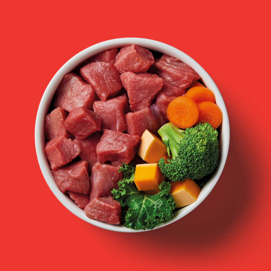 Bowl of Raw Boneless Beef 1kg with broccoli, carrots, and cheese cubes on a red background.