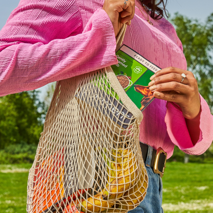 Person in pink sweater with a net bag of Senior Gently Steamed Turkey & Salmon 395g while shopping in the sunny park.