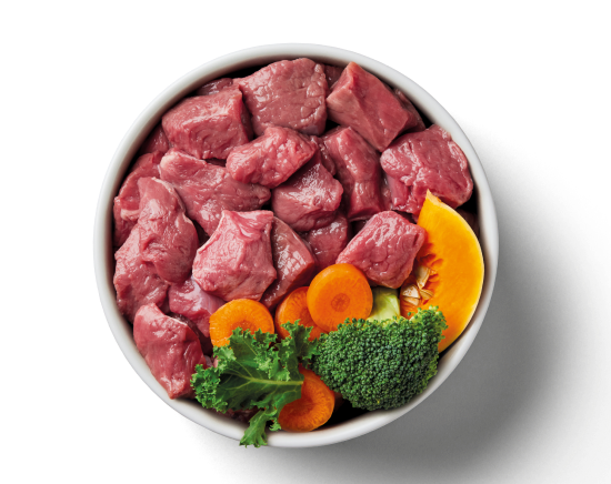 A bowl of raw meat chunks with broccoli, carrot slices, kale, and pumpkin pieces.