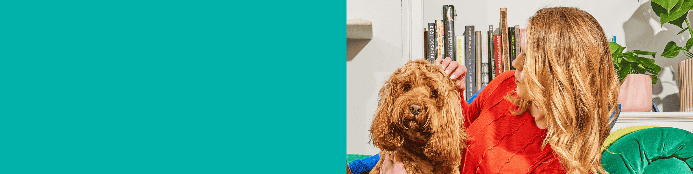 A woman pets a curly-haired dog while sitting on a sofa, with books and a plant in the background.