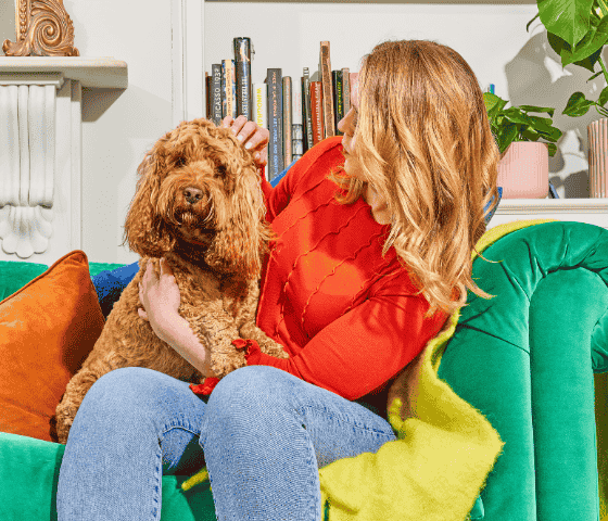 Woman in a red sweater sitting on a green sofa, holding a fluffy brown dog and looking at it.