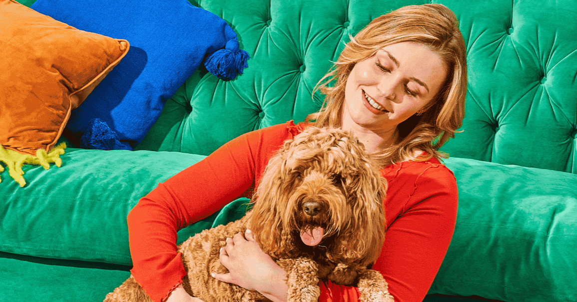 Woman in a red top smiling and holding a curly-haired dog on a green sofa with colorful pillows.