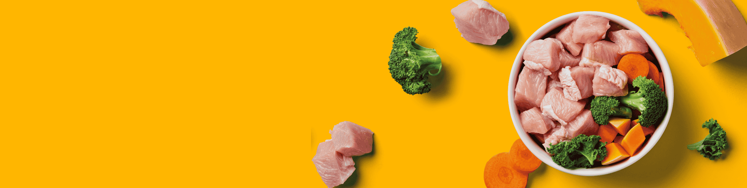 Bowl of raw chicken cubes, broccoli, and carrots on a bright yellow background with scattered ingredients.